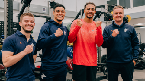 Ben is wearing a salmon top and has his fists clenched and is smiling at the camera. Alongside him are people from the sports centre dressed in navy sports attire also smiling and clenching their fists. Equipment can be seen behind them.