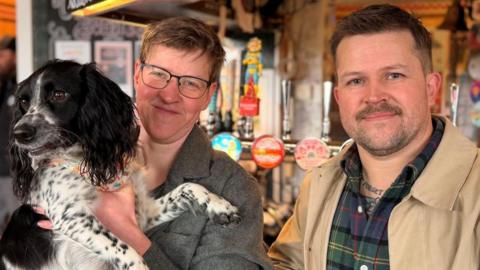 A man and a woman looking at the camera and smiling. The woman is holding a black and white Spaniel.