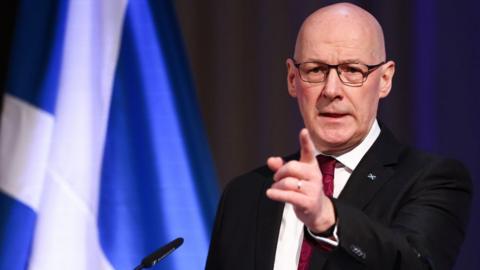 John Swinney, a bald man with glasses, photographed in a medium-close up shot. He is standing at a podium behind a microphone and in front of an illuminated saltire. He is wearing a dark suit, white shirt and red tie