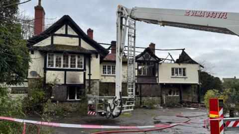 A derelict building. The roof is burned down. There is an aerial ladder platform in front of the building. There is fire tape in the front of the image. The sky is grey with grey clouds. There are bushes on either side of the building. 