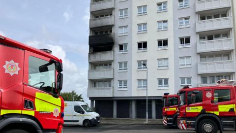 Three fire engines and a white van parked in front of block of flats, one of the flats has a burnt out balcony.