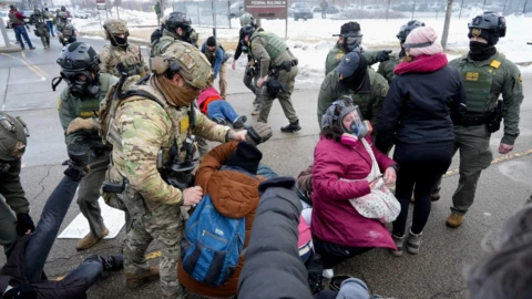 Border Patrol officers and Border Patrol tactical unit (BORDAC) officers surround protesters in Minneapolis. Two protesters in the foreground (a person in a brow jacket to the left; a woman in a pink parka and mask gas to the right) are kneeling to the ground; another protester in blue jacket stands in front of three officers. Another man is seen laying on the ground in the background