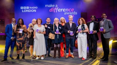 A row of a mixture of men and women who all hold a purple Make a Difference award whilst stood on stage. The backdrop is purple with "BBC Radio London" in white on the wall.