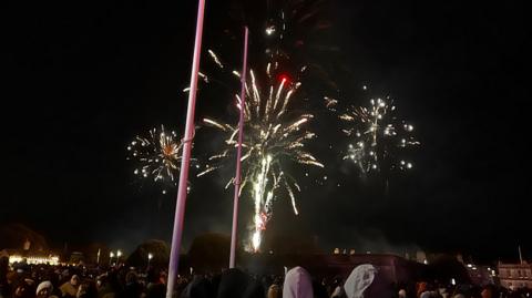 Gold and white fireworks in the sky on Plymouth Hoe. There are many people watching the fireworks. 