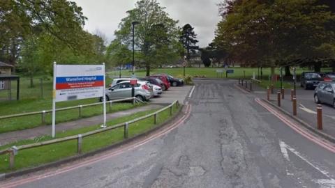 A google images photo of the main entrance to Warneford Hospital, showing a short road and a sign on the left with directions to different departments, and a car park on the right.