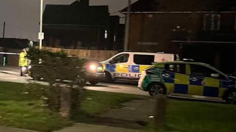 Stanway Road in Gloucester at night. There is a police van and car parked on either side of the road. A cordon is in place to the left of the photograph with two uniformed officers standing in front of it. Houses can be seen along the road.
