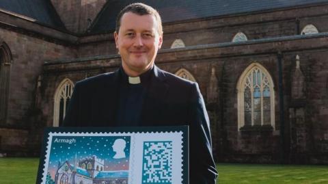 The Dean of Armagh, The Very Reverend Shane Forster, holding a small sign with the image of the stamp design on it - the stamp is an illustration of St Patrick's Cathedral with lots of children outside of it playing in the snow. The cathedral is covered in snow with the lights on and the sky has a dark sunset. The Dean is standing outside the cathedral while holding the stamp. 