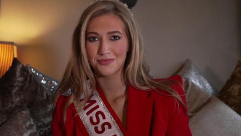 Tabitha sits on a couch surrounded by pillows. She has long blonde hair and wears a red suit, red and silver sash and silver crown.
