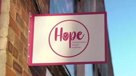 A pink and white sign on a brick wall with the Hope logo. It says "Hope, homelessness, hunger, hardship".