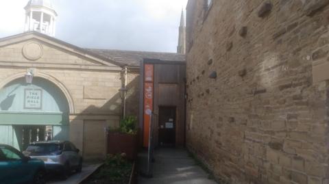 The front of a building with two entrances. The door on the left is arched and green with a sign that reads The Piece Hall. The door on the right has a sign up the left-hand side that reads Orangebox.