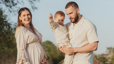 Chloe and her family all wear beige and stand outside in the sunshine with trees visible in the background as part of a professional family photoshoot. Chloe is visibly pregnant and holds her stomach smiling, while her partner holds their son.