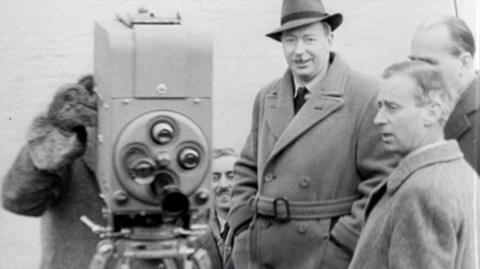 A group of men stand beside an old camera