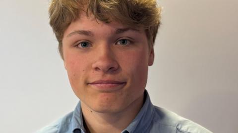 Gabriel Raimondo pictured in the BBC studio. He's wearing a blue shirt and a rosette in red, blue and white. He has wavy blonde hair and is smiling.