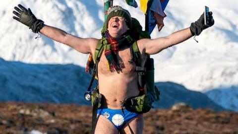 Speedo Mick Speedo Mick wearing his blue Speedos, a woolly hat, scarf, backback, gloves and nothing else, his face tilted up[wards to the sky with his eyes closed and a look of relief, against a background of snowy mountains in Glencoe, Scotland, in January 2023