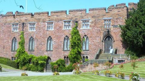 A grand old building made of red stone, with well-trimmed lawns to the front and sides