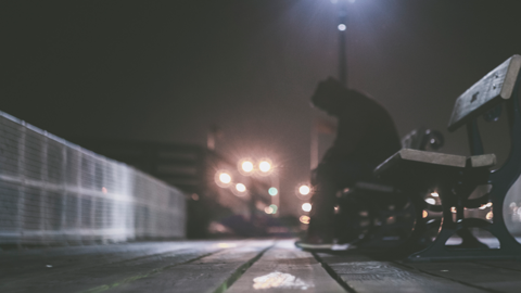 Person in silhouette sitting on a bench. Bright lights are in the background, slightly out of focus