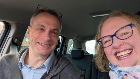 Man and women smiling at the camera sat in a car. He's got a seatbelt on and is wearing a grey jumper with a blue shirt. The women on the right has blonde hair and is wearing glasses and is taking the selfie.