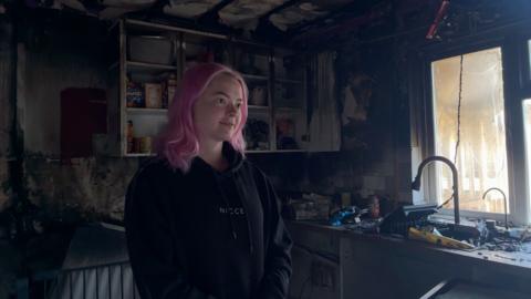 A woman in a black jacket with long pink hair inside a fire-damaged kitchen