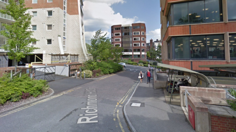 A screenshot from Google street view showing a narrow road with double yellow lines on each side. There's a block of flats on the left and an office block and a bike storage shed on the right.