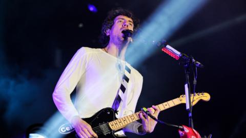 Gary Lightbody of Snow Patrol plays the guitar and sings on stage. He is wearing a white long-sleeved top.