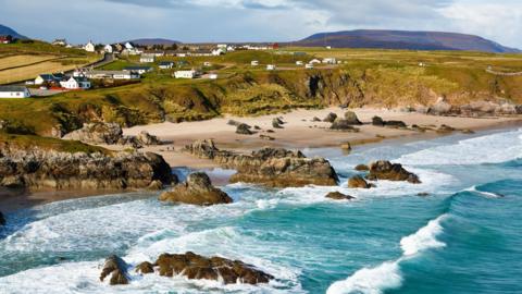 White walled properties dot an area of land near small cliffs above a beach. Waves roll in from the sea, which is a turquoise colour. There are rocky outcrops on the beach and in the sea.