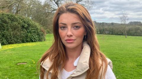 A woman with strawberry blonde hair stares at the camera. She's stood at a gate looking over a green field behind her, with hedges and trees in the background. The sky has grey clouds with a hint of blue skies in the distance. Lorna is wearing a white top, with a cream gillet over the top.