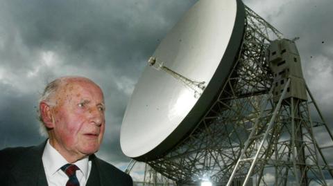 Sir Bernard Lovell, with thinning white hair, stands in front of the giant Jodrell Bank telescope.