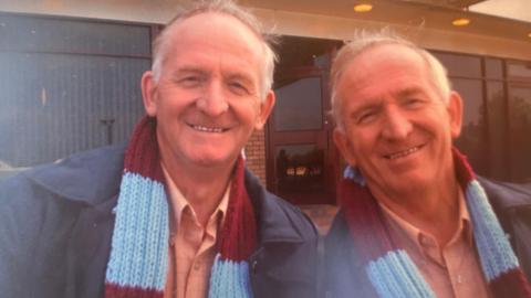 The twins are looking at the camera both smiling. They are wearing matching claret and blue scarves, pink button up shirts and blue jackets. Behind them is a building and door.
