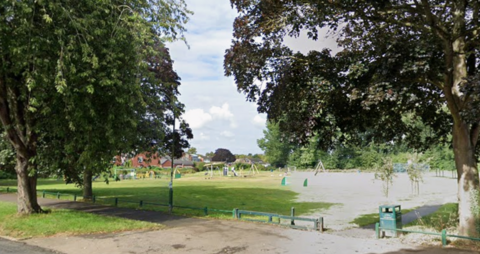 A view from Cubbington Road of The Holt. There are mature trees on either side of the picture and play equipment in the centre of the image.
