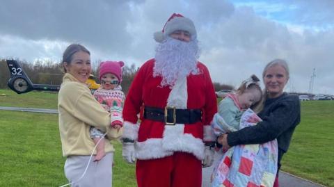 Jo with daughter Willow, aged 16 months, and Emma with Myla, aged two, greet Santa. Father Christmas is dressed in his traditional red and white outfit and the mothers are holding their children who have a blanket and a woolly hat to keep them warm. The group is standing outside.