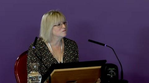 Joanne Hodson, who has shoulder length blonde hair, a fringe and glasses, sits at a witness table in front of a screen and a black microphone in front of a purple screen. 
