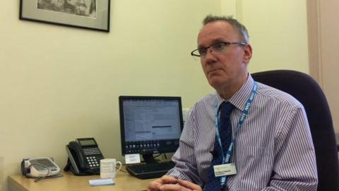 A doctor is sat at a desk which has with a computer and telephone and a mug on it. He is wearing a purple and white striped shirt, with a blue NHS lanyard and name badge.