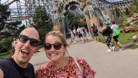 Dean Stokes - a man with no hair smiles into the camera. He is wearing sunglasses and next to a woman, also wearing sunglasses. She is smiling, wearing a pink and black dress and has blonde hair.