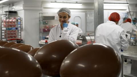 Nina gazes at some big easter eggs in a factory. She's wearing blue gloves, a hair net and a white coat