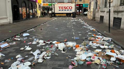 A white Tesco-branded lorry is the focal point of the photo, with a litter-covered road leading up to the lorry. Litter includes food boxes, plastic cups and bottles