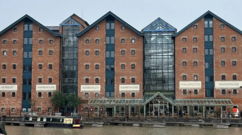 Herbert, Kimberley and Philpotts warehouses pictured from across the river. They are large brick building which say "Herbert Warehouse" "Kimberley Warehouse" and "Philpotts Warehouse" on them. There is a large glass entrance between the Kimberley and Philpotts buildings and two canal boats can be seen on the water.