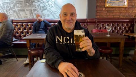 Simon Berry sits at a wooden table in a traditional pub, wearing a dark hoodie and holding a pint of pale beer. The cosy setting features leather bench seating, wooden floors, brick walls, and nearby patrons