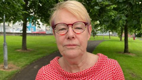 A woman with short blonde hair and purple highlights is wearing multicoloured framed glasses and is looking into the camera. She is wearing a red and white patterned top. She is standing on a path in a green area surrounded by trees.