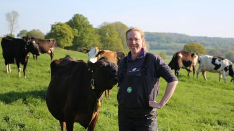 Dairy farmer Lorna Burdge stands in a field wearing overalls, with cows behind her