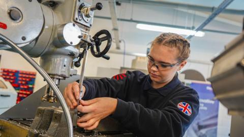 A woman wearing a jumper which has a union flag on the arm, is seen using a tool to drill downwards into some material. She is looking at her job.