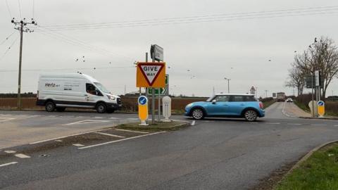 Looking across a four-way crossroads in a rural area as a white van and a blue car pass under a grey sky. A give-way sign and other signs stand in the centre of the picture. A lorry and a second car are in the distance. The land is flat with a brown field and bare trees, with birds sitting above on power lines.