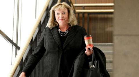 Marilyn Gladu walks down a staircase in a black suit with a silver necklace. She is holding a red takeout coffee cup in her hand as well as a black purse.