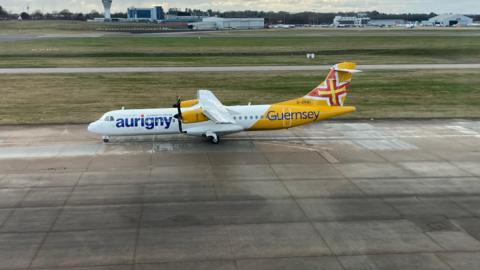 An Aurigny aircraft on an airport runway. The plane is yellow and white and has two propellers.