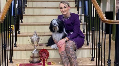 Paige Le Moignan and Mika sit on a set of tiled stairs with black metal banisters either side of them. Paige is wearing a purple-above knee length dress with diamantes covering the neckline and the hem. Mika is black and white in colour and has hair covering her eyes. They are sat next to their trophy which is silver with large handles and is about 20 inches tall. Next to the trophy is a red and gold rosette. 