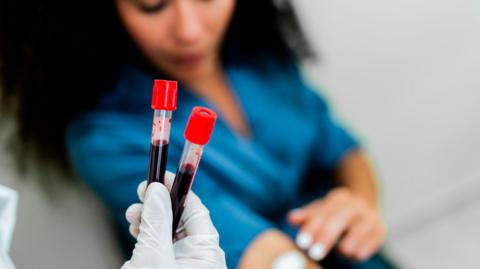 A woman puts a piece of cotton wool onto her arm after giving blood. In the foreground of the picture, a healthcare professional holds two samples of blood in a gloved hand.