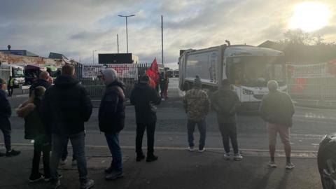 A group of people are standing on a pavement opposite a bin depot. The sun is rising in the cloudy sky. There is a large bin lorry driving out of a gate. There are red flags on the metal fences surrounding the depot exit
