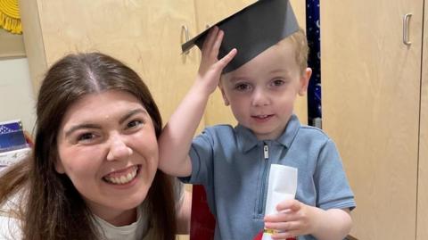 Conor holds his little black graduation mortar board hat on his head. Cupboards are behind him in a nursery.