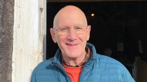 Mr Harrison is standing in a doorway. The background appears to be a dimly lit interior space. The doorway frame on the left side is made of light-coloured, weathered wood. He is wearing a teal quilted jacket over an orange top. He is smiling at the camera and also wearing glasses.