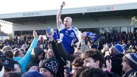 Macclesfield's Josh Kay raises his arm in the air celebrating while surrounded by fans on the pitch. His mouth is open and he looks to be cheering. He appears to be on the shoulders of a team-mate and raised above the surrounding fans.