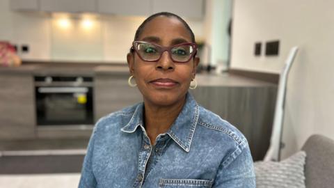 Patricia Ogunfeibo, wearing shaped dark red coloured glasses, hooped earrings and a denim collared shirt, is sitting with a kitchen worktop in the background behind her